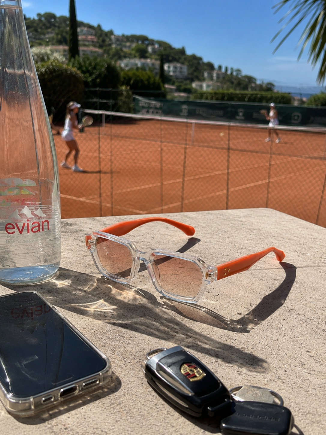 Clear Mareframes sunglasses with orange temples on a sunlit table by a clay tennis court